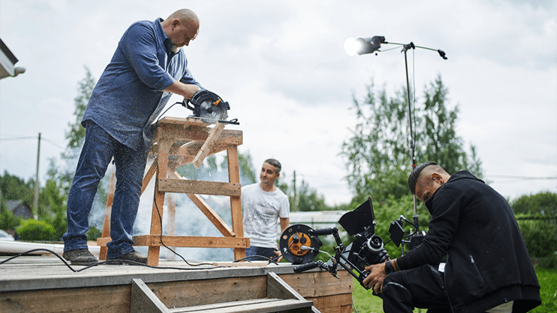 Оператор снимает как мужчина пилит циркулярной пилой